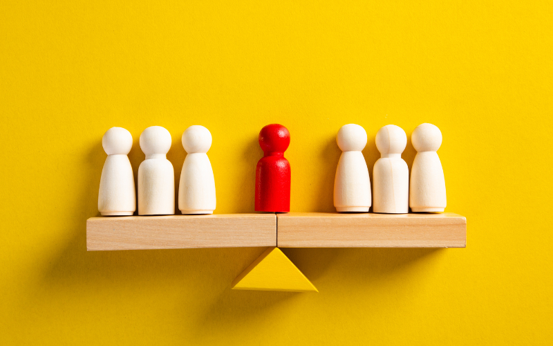 Two groups of figurines are balancing on either end of of a balance with an red colored figurine representing an arbitrator in the middle.